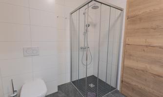 A modern bathroom with a glass shower cabin and a white toilet. The walls are tiled in light colors and the floor is dark gray.