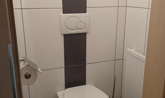 A small bathroom with a modern design. It contains a toilet, a roll of toilet paper, and a waste bin.