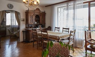 A bright dining room with a large wooden table and chairs. The walls are adorned with windows and a decorative showcase.