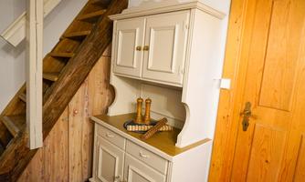 A bright wooden cabinet stands in a room next to a staircase. Inside are spice mills and a decorative tray.