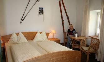 A cozy bedroom with a wooden bed and two pillows. In the room are skis and a table with a person who is relaxing and enjoying a drink.