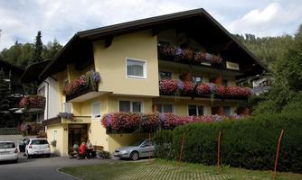 A charming building with a yellow facade and colorful flowers on the balconies. In the foreground, some cars are parked and a cozy seating area is visible.