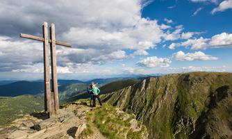 A couple poses at the summit of a mountain next to a wooden cross. The sky is blue with some clouds, and the surrounding mountains are green and impressive.