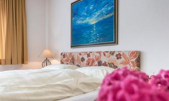 A cozy bedroom with a large bed and a colorful headboard. On the wall hangs a colorful painting and on the nightstand stands a lamp.