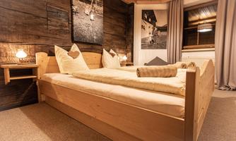 A cozy bedroom with a wooden bed and lovingly designed pillows. The walls are made of wood, and large windows let in plenty of light.
