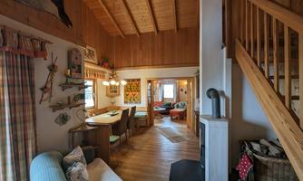 A cozy living room with a wooden ceiling and a dining area. On the right side, a staircase leads upstairs and decorative elements hang on the wall.