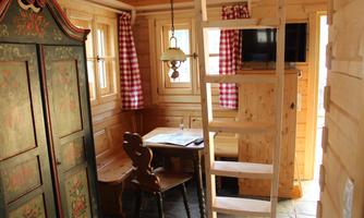 A cozy wooden interior with a table, chairs, and a cabinet. A ladder leads to an upper area, and windows with checked curtains let in plenty of light.