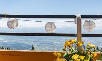 A view from a balcony with paper lanterns and yellow flowers. In the background, mountain landscapes and a blue sky are visible.