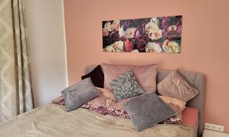 A cozy bedroom with a bed decorated with several pillows. A picture with rose motifs hangs on the wall.