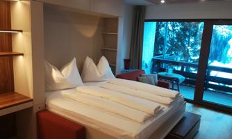 A cozy bedroom with a large bed and white pillows. Large windows offer a view of the balcony and the surroundings.