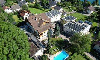 An airy view of a building with solar panels and a blue pool. Surrounded by green landscapes and other residential houses.