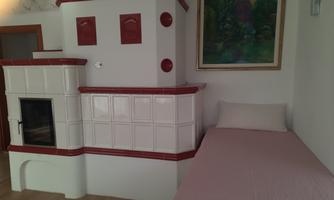 A cozy room with a tiled stove in white and red. On the right is a simple bed with a light blanket.