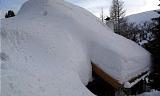 A house is buried under a thick layer of snow. In the background, some trees are visible.