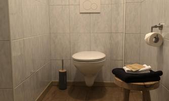 A modern bathroom with white tiles and a toilet bowl. Towels are laid on a wooden chair and a small plant is sitting in the window.