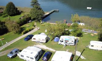 A campsite by the water with several caravans and cars. In the background, there are trees and a harbor.