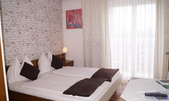 A cozy bedroom with a double bed and light curtains. An artwork hangs on the wall and a table lamp provides atmospheric light.