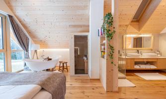 A modern room with wooden paneling and large windows. It features a bathtub, a washbasin, and a shower, surrounded by natural light.