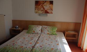 A simple bedroom with a double bed and colorful bedding. A decorative picture hangs on the wall and there are wooden shelves next to the bed.