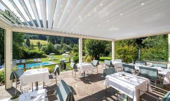 An elegant restaurant with tables placed outside. In the background, there is a garden and a pool, surrounded by nature.