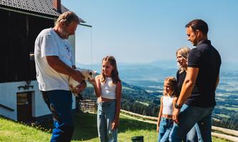 A group of people is standing outside on a meadow overlooking a picturesque landscape. A man is holding a small dog while the others are conversing amicably with one another.