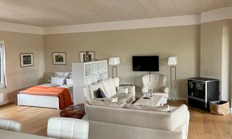A modern furnished living room with comfortable sofas, a television, and a bedroom in the background. The room has large windows and a warm wooden floor.