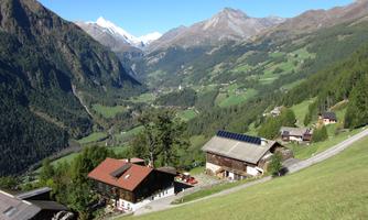 A picturesque view of a high valley with green meadows and several chalets. In the background, mountains with snow-capped peaks are visible.
