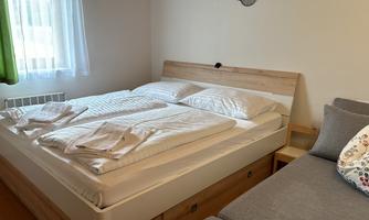 A cozy bedroom with a large double bed and a comfortable sofa. The windows let in plenty of daylight and the walls are painted in light colors.