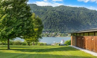 A peaceful landscape with a green lawn and a view of a lake. In the background, gentle hills and a few small buildings can be seen.