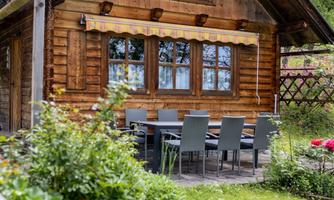 A charming wooden house surrounded by a garden. There is an outdoor dining table with chairs and blooming plants.
