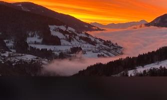 A breathtaking sunset over the mountains, with an orange sky and fog filling the valleys. The snowy slopes give the landscape a picturesque atmosphere.