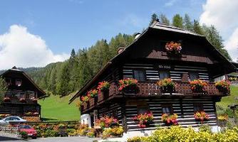 Two traditional wooden houses surrounded by colorful flowers and a green landscape. The sky is clear with some clouds.