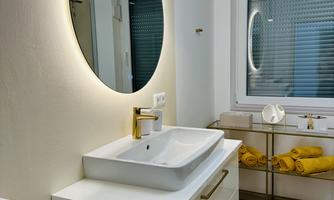 A modern bathroom with an elegant sink and a round mirror. There is wooden flooring and yellow towels on a shelf.