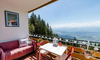 A cozy terrace with a red sofa and a small table. In the background, an impressive mountain landscape stretches out under a clear blue sky.