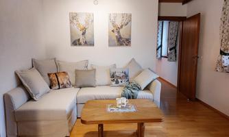 A cozy living room with a bright sofa and decorative cushions. On the wall, two pictures of deer hang, and the wooden window lets in plenty of light.