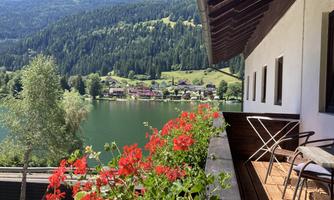 A beautiful view of a calm lake, surrounded by green mountains. Blooming flowers adorn the terrace of a house.