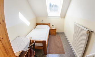 A simple bedroom with a single bed, a table, and a chair. It has daylight through a window and a warm, bright shade.