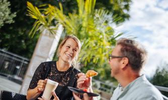 A cheerful couple is sitting outdoors and enjoying coffee and pastries. Behind them, plants and a blue sky are visible.
