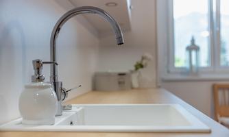 A modern kitchen sink made of white material with an elegant faucet. In the background, windows and a cozy dining area are visible.