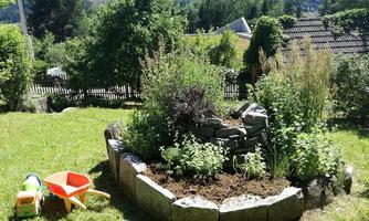 A lovely garden with a planted raised bed and colorful toys. In the background, there are trees and a gentle hill.