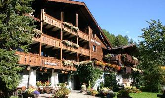 A charming wooden house with multiple balconies and colorful flowers. The surroundings are green and inviting, perfect for a relaxing stay.