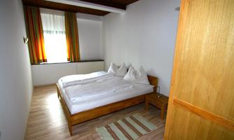 A simple, modern bedroom with a double bed and wooden furniture. The large windows provide plenty of daylight.