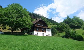 A farmhouse surrounded by green meadows and trees. The sky is blue with white clouds.