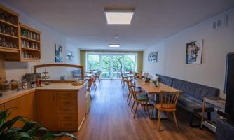 A modern dining room with wooden tables and chairs.  
Large windows offer a view of greenery.
