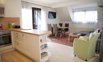 A modern, bright apartment with an open kitchen and a cozy living room. The dining area is furnished with a table and chairs, and large windows provide plenty of daylight.