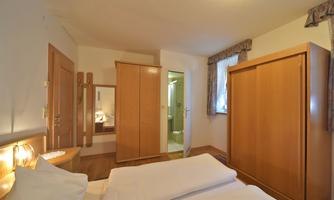 A bright bedroom with a double bed and wooden wardrobes. The bathroom is accessible through a door.