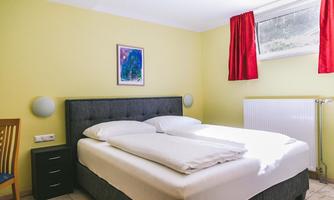 An inviting bedroom with a large double bed and colorful curtains. The walls are painted in a friendly yellow tone and a window lets in daylight.