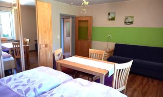 A cozy room with a table and chairs, a sofa, and two beds. The walls are partially painted green, and there are windows that provide natural light.