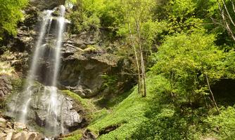 A picturesque waterfall cascading over large rocks into a green surroundings. Lush trees and bushes surround the scene, creating a peaceful atmosphere.