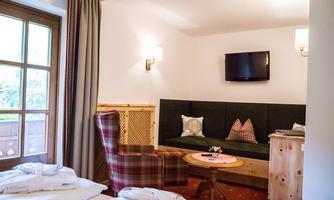 A cozy hotel room with a comfortable armchair, a couch, and a table. A television is mounted on the wall and a window lets in daylight.