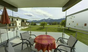 A balcony with a round table and chairs. In the background, green meadows and mountains can be seen.